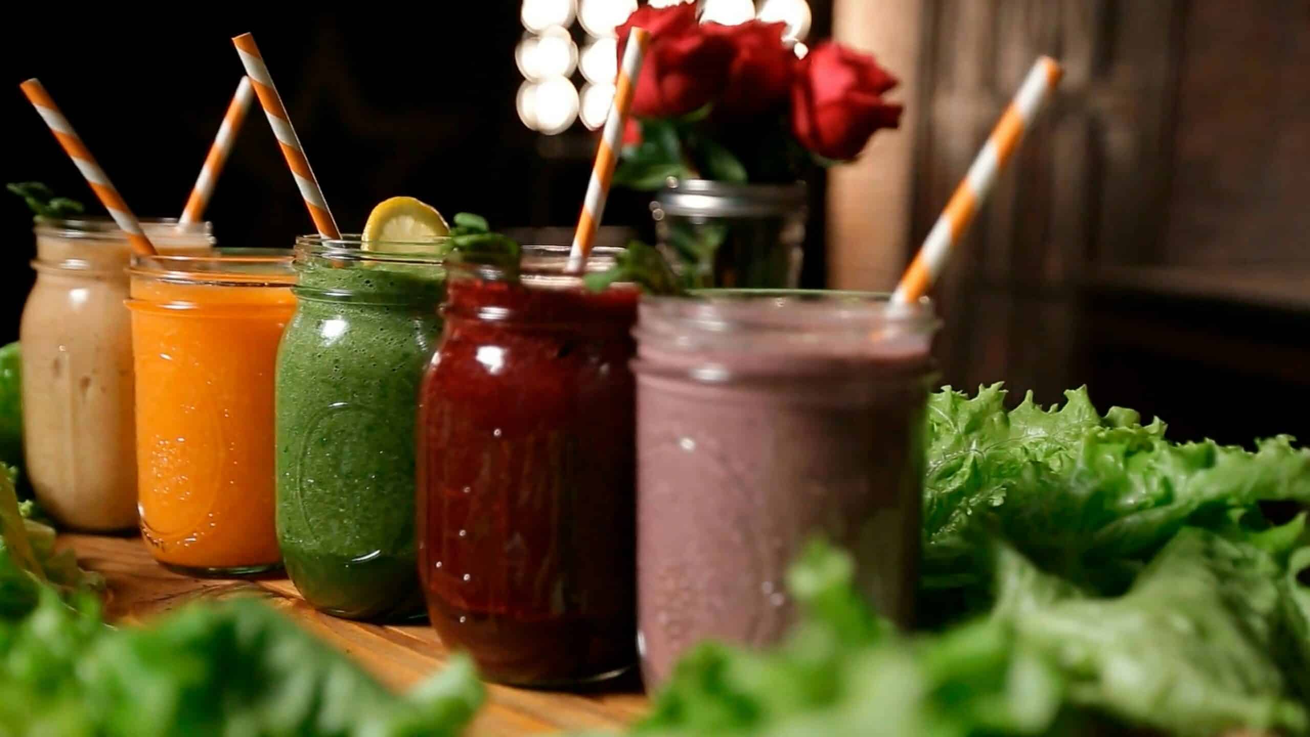 Cinq smoothies aux couleurs variées, préparés à base de fruits et de légumes frais, présentés dans des bocaux en verre avec des pailles.